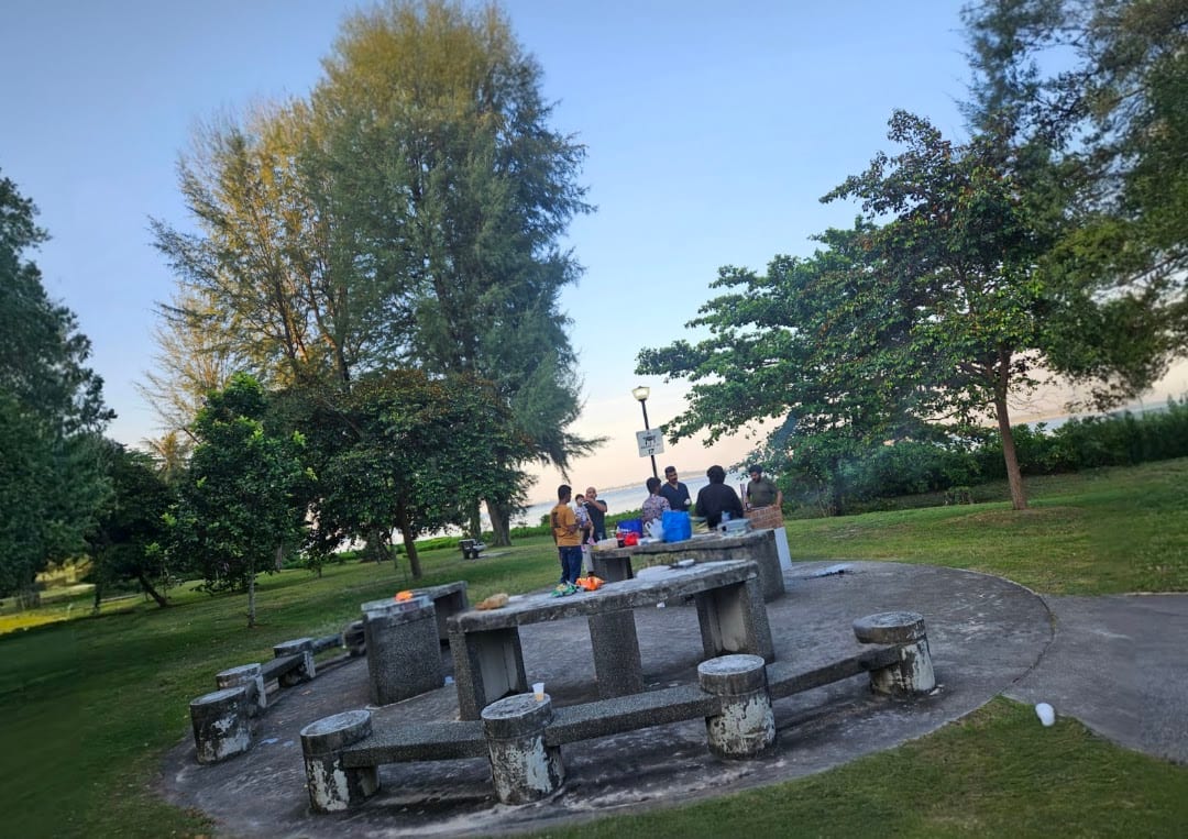 A concrete barbecue pit with tables and stools at Changi Beach Park, with a group grilling at dusk